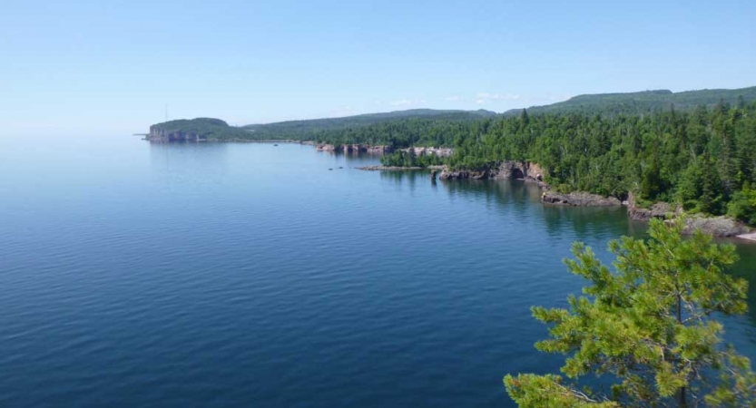 The rocky and tree-lined shore of Lake Superior frames the calm, blue water of the lake. 